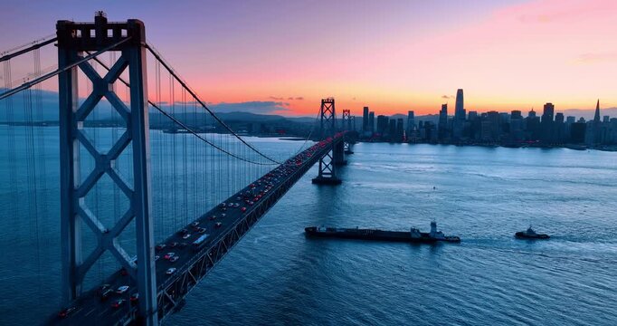 Flying along the Oakland Bay Bridge with hectic traffic on. An empty container ship moves under the bridge. Skyline of San Francisco, California, USA at sunset in the backdrop.