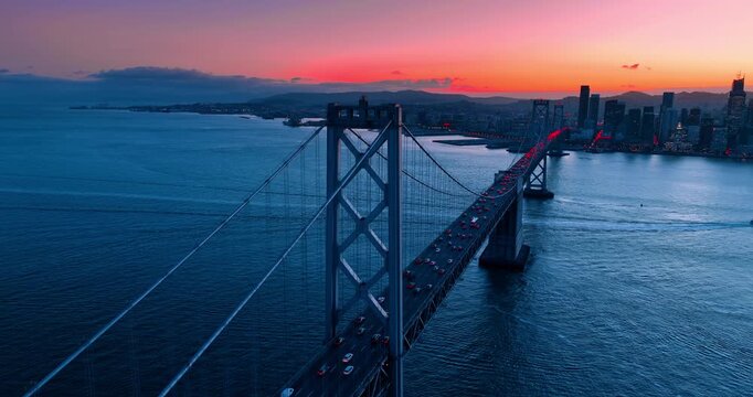 Numerous cars move in the hectic traffic by the Oakland Bay Bridge at sunset time. San Francisco skyline against pink sky in the backdrop.