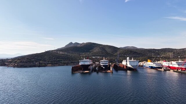 Old decommissioned ferry ships and cargo vessels moored in bay with green hills in background, Greece maritime scrapyard and ship breaking facility
