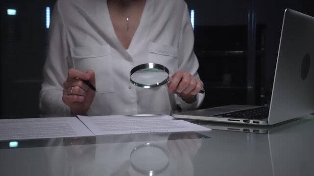 Businesswoman conducting detailed audit checking financial spreadsheets using magnifying glass with laptop and documents on desk. Audit and taxes concept