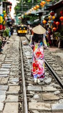Woman walking along Hanoi train street with colorful Vietnamese lanterns and vintage steam locomotive approaching between narrow houses in Vietnam.