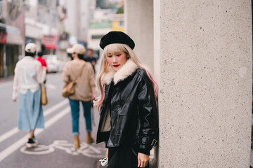 Obraz premium Young woman with blonde hair wearing black leather jacket and beret standing on city street with blurred pedestrians walking in background, urban fashion style