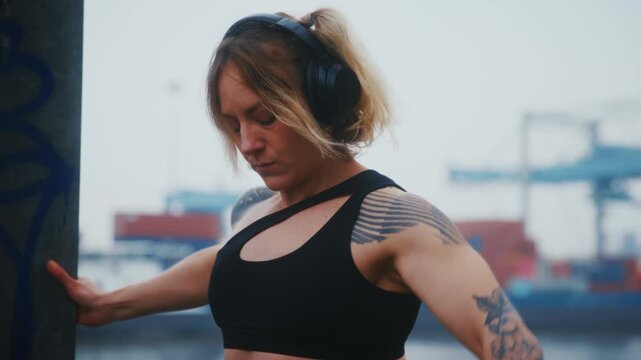 Tattooed Athletic Woman Doing Dumbbell Front Raise with Headphones at Industrial Harbor Waterfront