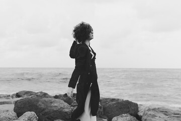 Woman in a long coat stands on a rocky shore by the sea, wind in her hair, looking toward the horizon in a moody monochrome landscape © SHOTPRIME STUDIO