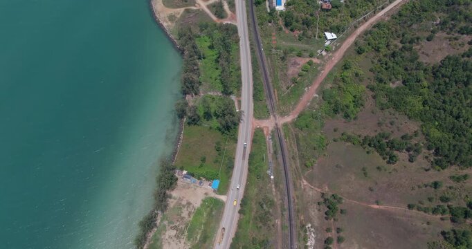 Top down aerial road with cars and trucks, parallel railway tracks, turquoise ocean, and green lands
