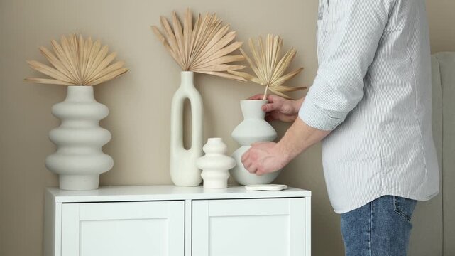 Man putting vase with dried palm leaf onto cabinet near beige wall indoors, closeup