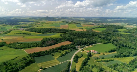Fototapeta premium Green fields and hills stretch across the countryside with a winding road and small houses below in summer light