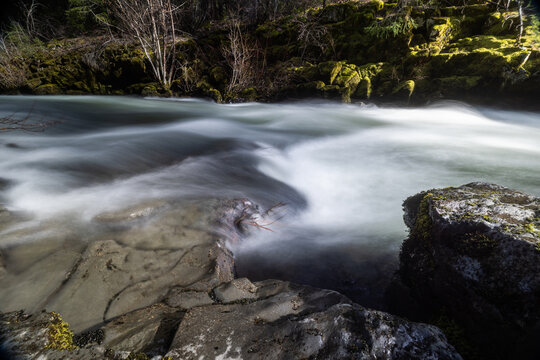 Rogue River above Union Creek, Oregon rushes over jagged stones. Mossy banks glow in golden sunlight. Nature's force moves through rocky terrain.