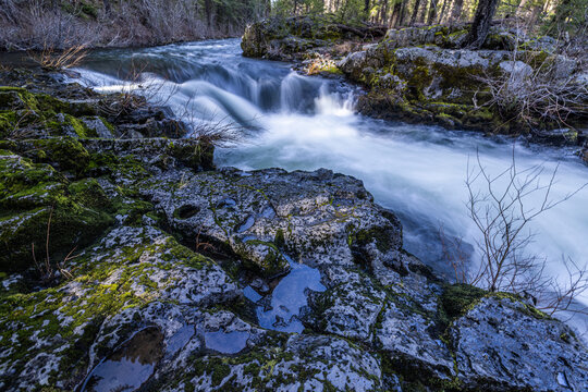 Rogue River above Union Creek, Oregon rushes over jagged stones. Mossy banks glow in golden sunlight. Nature's force moves through rocky terrain.