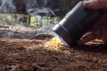 Obraz premium Powerful flashlight starts a fire on kindling. Fingers grip flashlight, steady, focused. Fire crackles low beneath forest floor. Dirt, twigs, and leaves surround glowing flame