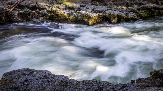 Rogue River above Union Creek, Oregon rushes over jagged stones. Mossy banks glow in golden sunlight. Nature's force moves through rocky terrain.