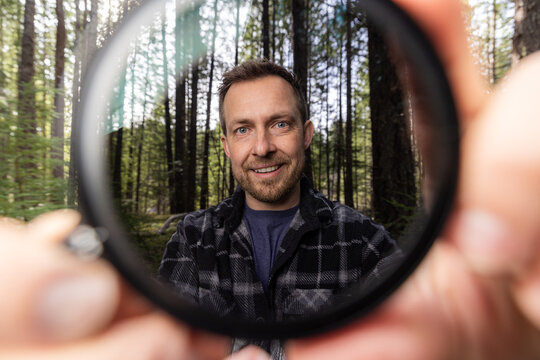 Man smiles through a neutral density filter for the camera . Green trees blur behind his face. Natural light filters through the canopy. He holds the camera filter near the lens.