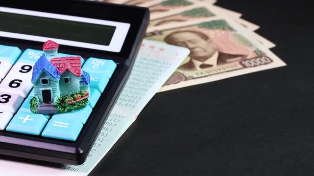 黒背景で回転する一万円札と通帳と電卓とミニチュアハウス