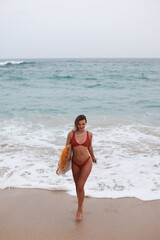 Fototapeta premium Passionate Surfer Exiting Ocean Shore Carrying Cheerful Yellow Surfboard And Wearing Lively Red Bikini