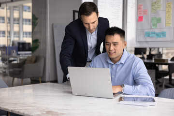 Obraz premium Workplace guidance and assistance. Experienced mid adult businessman in formal suit stand close to seated male intern trainee review laptop screen give consultation expertise feedback help with work