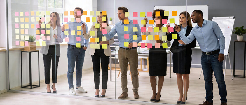 Panoramic view of a diverse professional team collaborating in a modern office, brainstorming with colorful sticky notes on a glass wall for agile project management and creative strategy.