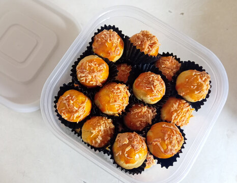 Close-up of homemade mini cheese ball pastries topped with shredded cheese, arranged in black paper cups inside a transparent plastic container on a white table. A popular sweet snack often served for
