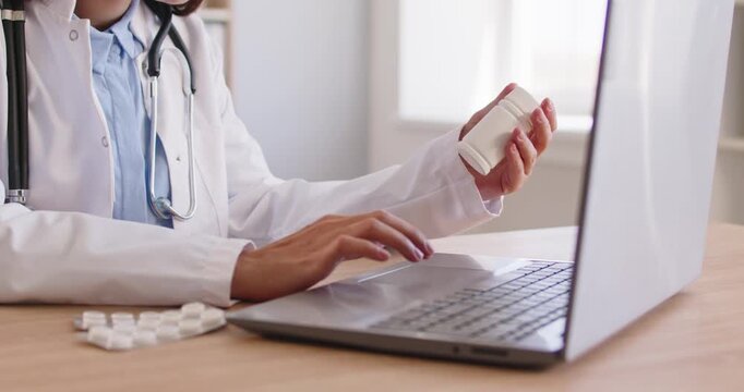 Doctor checking online medicine or vitamin pill instruction, side effects and dosage, sitting at laptop by window in clinic. Healthcare professional holding bottle in search of prescription guidance