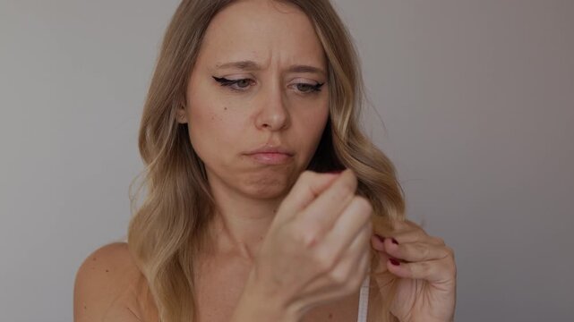 Young blonde woman examining dry damaged hair ends with concerned expression. Hair care, split ends and unhealthy hair concept