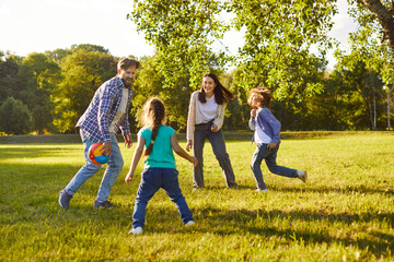 Cheerful parents and two children playing ball together on green lawn in park on sunny day. Happy smiling family members enjoying fun outdoor activity, bonding and spending quality time in nature.