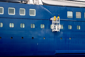 Naklejka premium Two deck hands in yellow rainsuits washing the side of a large blue ship