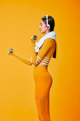 Obraz premium Female athlete in bright orange activewear lifts dumbbells in a bold studio, wearing headphones and a towel around her neck, showcasing strength, focus, and energetic fitness training in a vivid