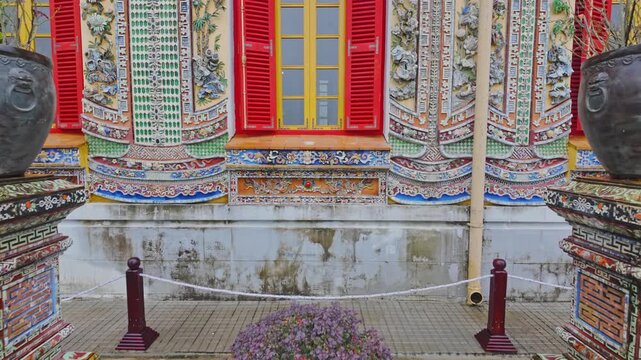 Two large decorative pots sit on ornate pedestals in front of a building with colorful windows and walls in  the Hue Imperial City, Hue, Vietnam. The pots add to the beauty of the