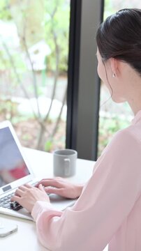 デスクで座るパソコンの仕事をするピンクの服を着た綺麗な女性の縦動画　キャリアウーマンなどの働く女性イメージ　顔無しの後ろ姿