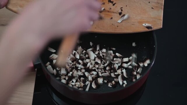Chopped mushrooms are transferred from a wooden board to a frying pan over heat. The hands work quickly to ensure even cooking.