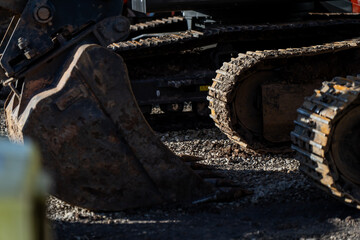 Fototapeta premium Excavator bucket beside steel crawler tracks on gravel ground, highlighting earthmoving equipment wear, contractor readiness and site operations