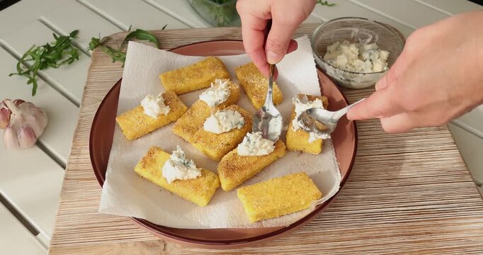 Anonymous person spoons gorganzola cheese onto pieces of crispy fried polenta on plate. Traditional Italian appetizer. Seasonal farm-to-table produce.