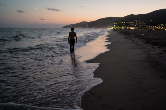 Young blond muscular man walking barefoot on beach at dusk