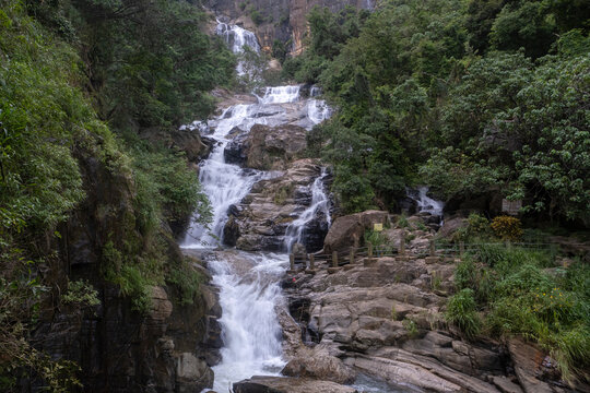 Panoramic view of Ravana Falls in Ella, Sri Lanka. Majestic wide waterfall cascading down rocky layers surrounded by lush tropical greenery and steep cliffs.