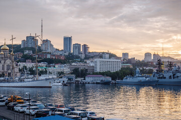 Modern warships and classic sailboats are docked in vladivostok harbor at sunset, with the cityscape rising behind them © Довидович Михаил