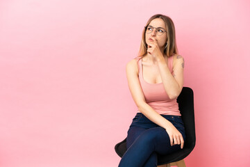 Young woman sitting on a chair over isolated pink background having doubts and with confuse face...