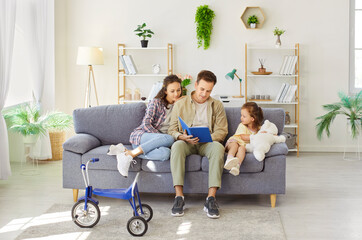 Fototapeta premium Parents reading fairy tale book with daughter at home together. Father sitting between little kid and woman on sofa, holding book to read children literature to listening family in living room