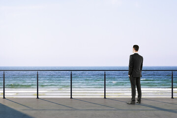 Fototapeta premium Businessman standing on seaside terrace looking at calm ocean horizon under bright sky in peaceful contemplative moment today.