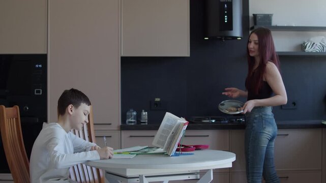 Schoolboy doing homework at kitchen table while mother serving food in kitchen