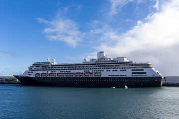 Large cruise ship docked at harbor with calm blue waters and clear skies, showcasing multiple decks and amenities along the side of the vessel