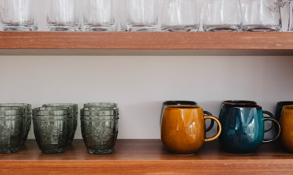 Glasses and coffee mugs sitting on a timber shelf.