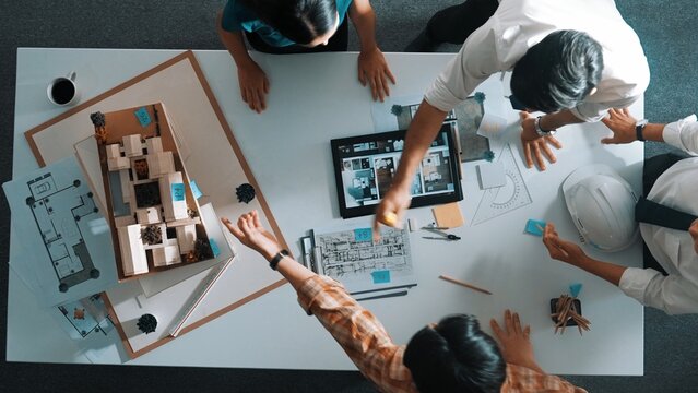 Top down view of architect engineer team discussing and point at tablet display construction at table with blueprint and house model at meeting. Diverse designer planning design house. Alimentation.