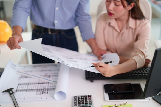 Asian architects engineers adult male female review blueprints design construction project plans collaborate office meeting teamwork discuss ideas solution focus work