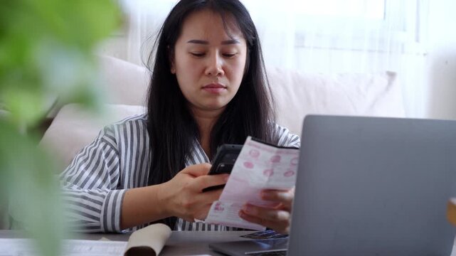 Focused Asian woman using a smartphone to scan a QR code on a utility bill for mobile payment while working on a laptop at home. Modern digital banking and financial management concept.