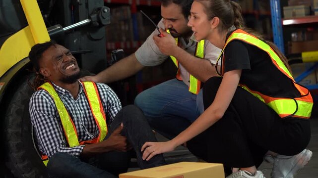 Industrial accident in warehouse. African American man worker lying on floor with leg injury after forklift incident. Caucasian coworkers providing first aid and calling for help. Workplace safety.