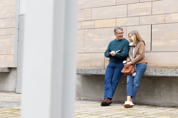 Fototapeta premium Senior couple leaning against concrete ledge in plaza holding book and coffee cup, copy space
