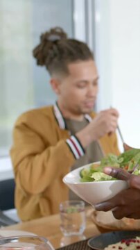 Vertical video: Offering bowl offframe urging man taking salad with fork at table in bomber jacket