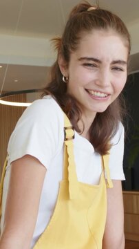 Vertical video: Smiling woman in yellow overalls seeing camera, leaning and laughing in modern room