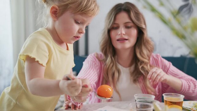 Young girl paints Easter egg with mother. Mother smiles warmly at child. Child focuses intently on coloring egg. Bright, cheerful kitchen setting. Perfect for Easter, family, or DIY craft themes
