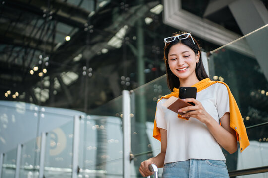 Young asian woman traveler using smartphone for checking flight information check the routes trip while waiting with her luggage at the airport terminal. Traveller woman passport and baggage luggage