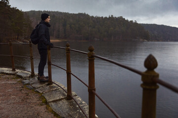 A man in a black beanie and jacket stands by a rusted railing on a stone path, gazing across a calm lake toward a dense, misty forest under a moody, overcast sky. Perfect for winter travel themes. © Francis